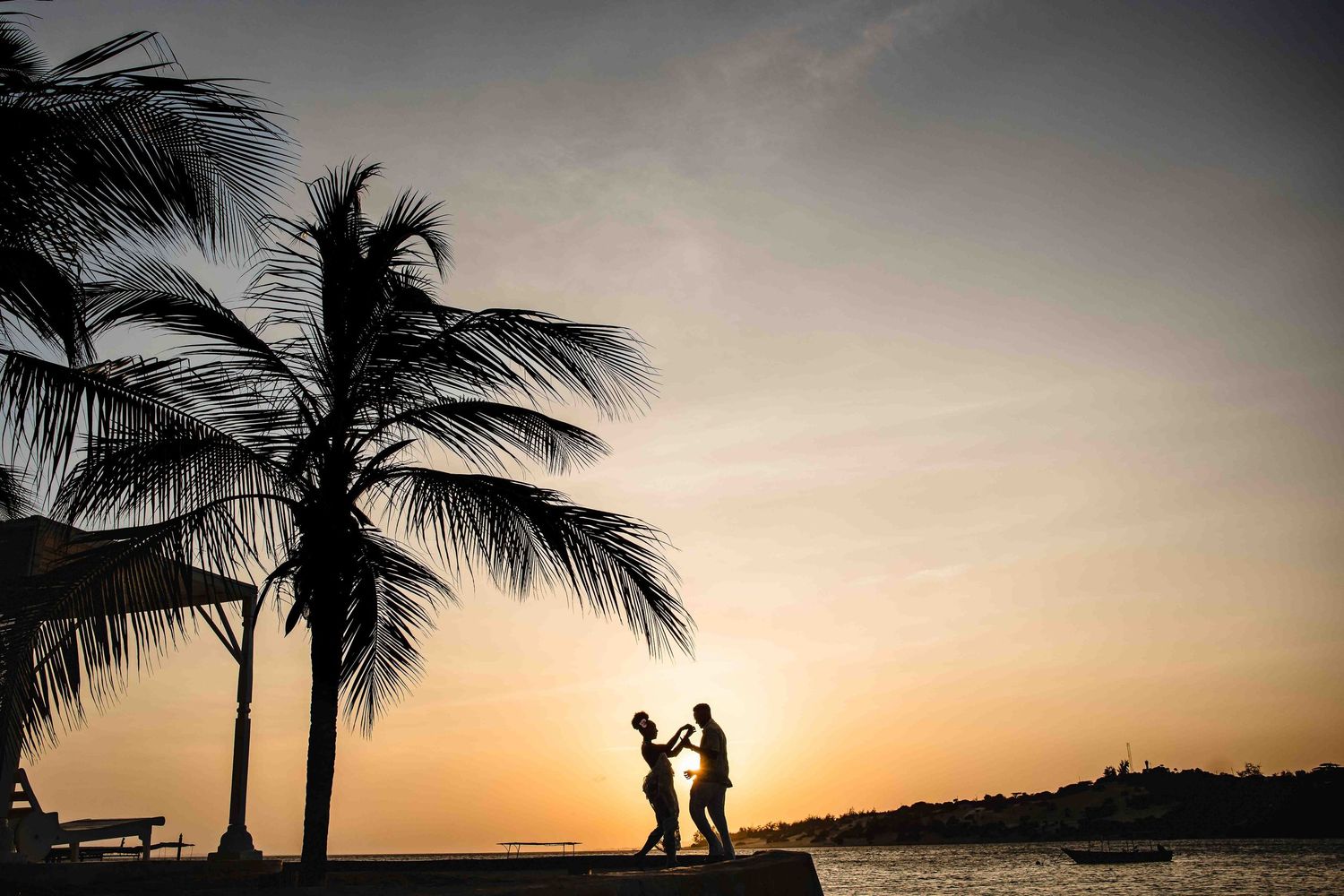 Palm trees sway against a vibrant orange sunset sky as silhouetted figures walk along the beach at dusk.