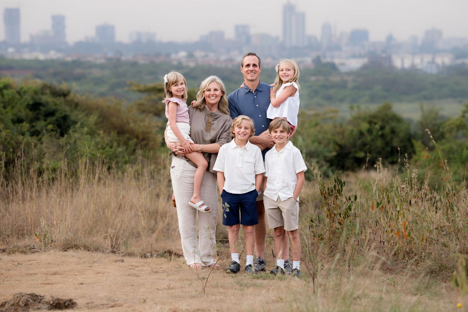 A family wearing coordinated white and blue outfits poses together on a grassy hilltop during sunset.