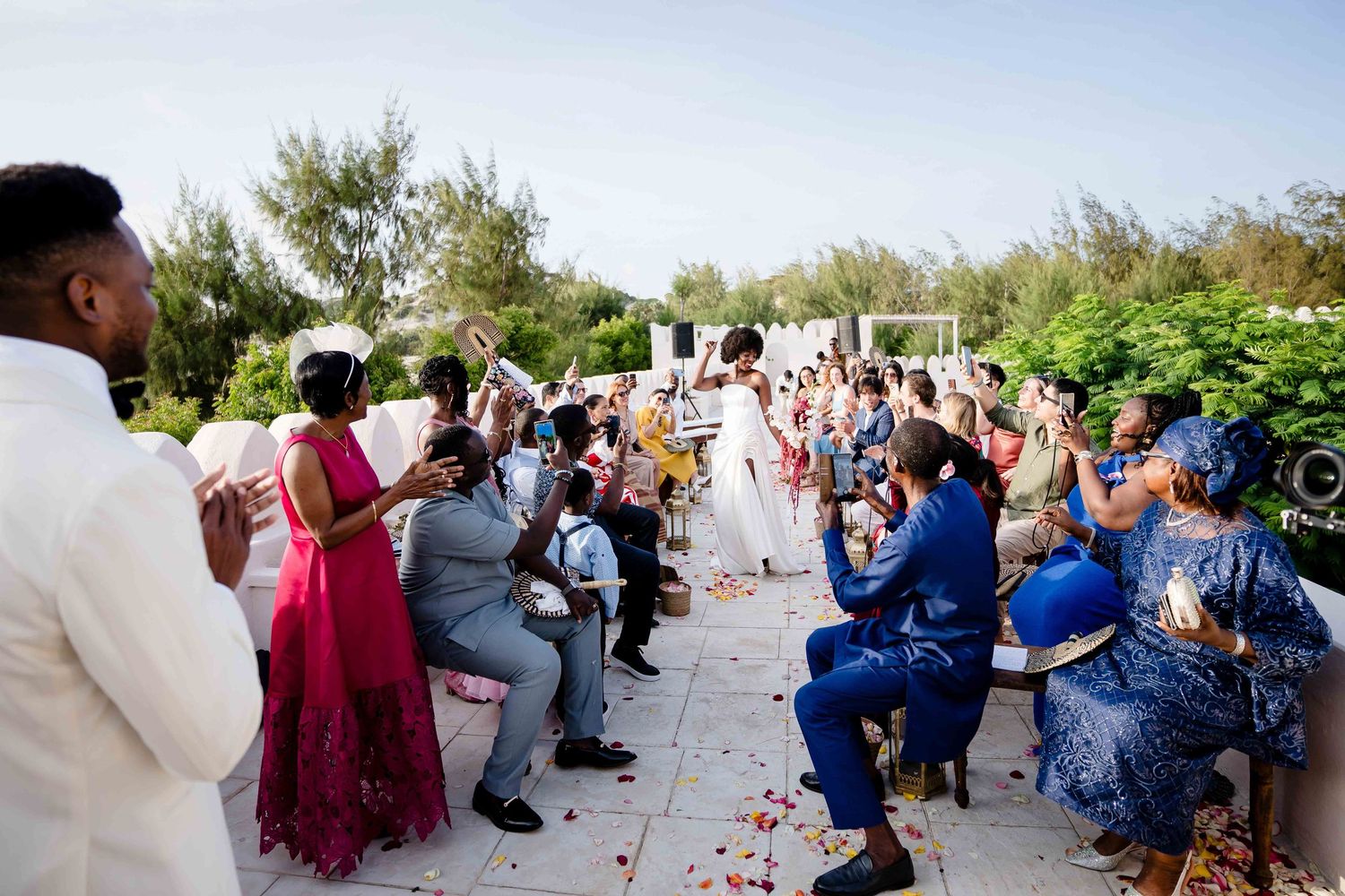 Wedding guests dance and celebrate at an outdoor garden reception with rose petals scattered on the ground.