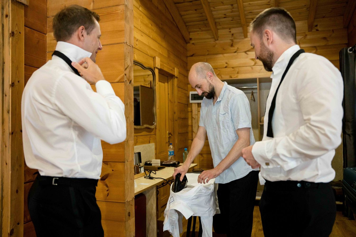 Three men in formal attire gather around a table during wedding preparations in a rustic wooden cabin.