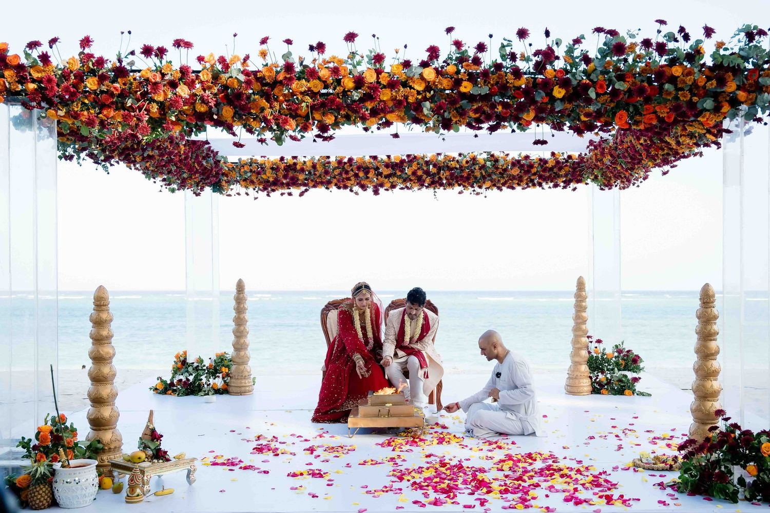 A beachfront wedding ceremony under a white canopy adorned with cascading orange and red flowers by the ocean.