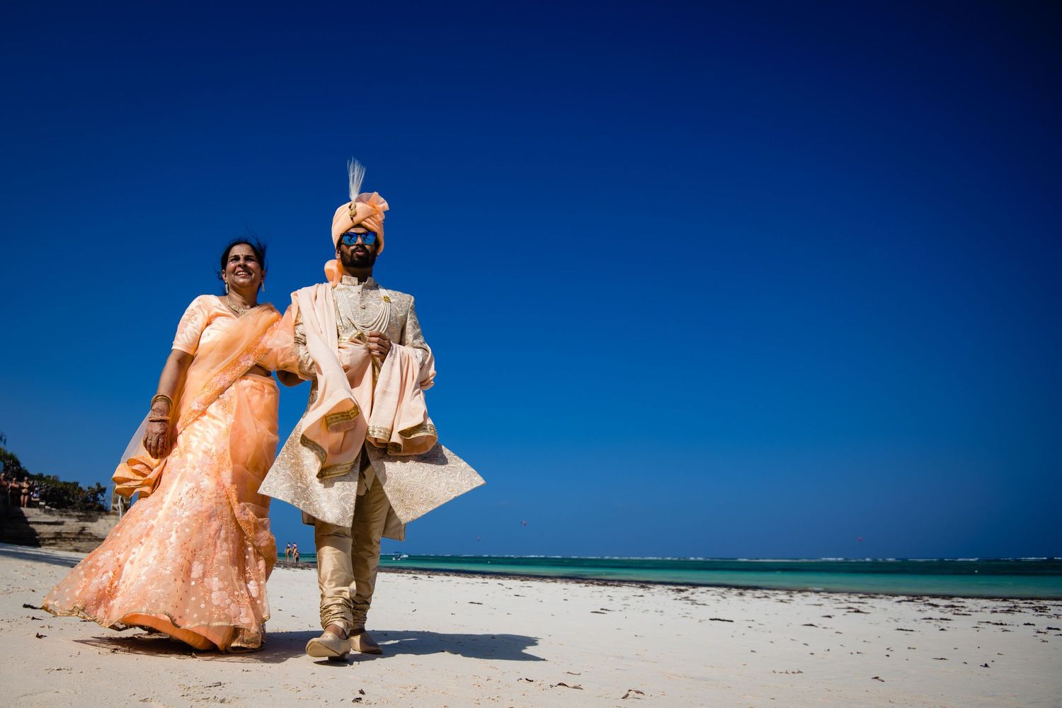 Couple in formal attire walking along a sunny beach with blue ocean backdrop.