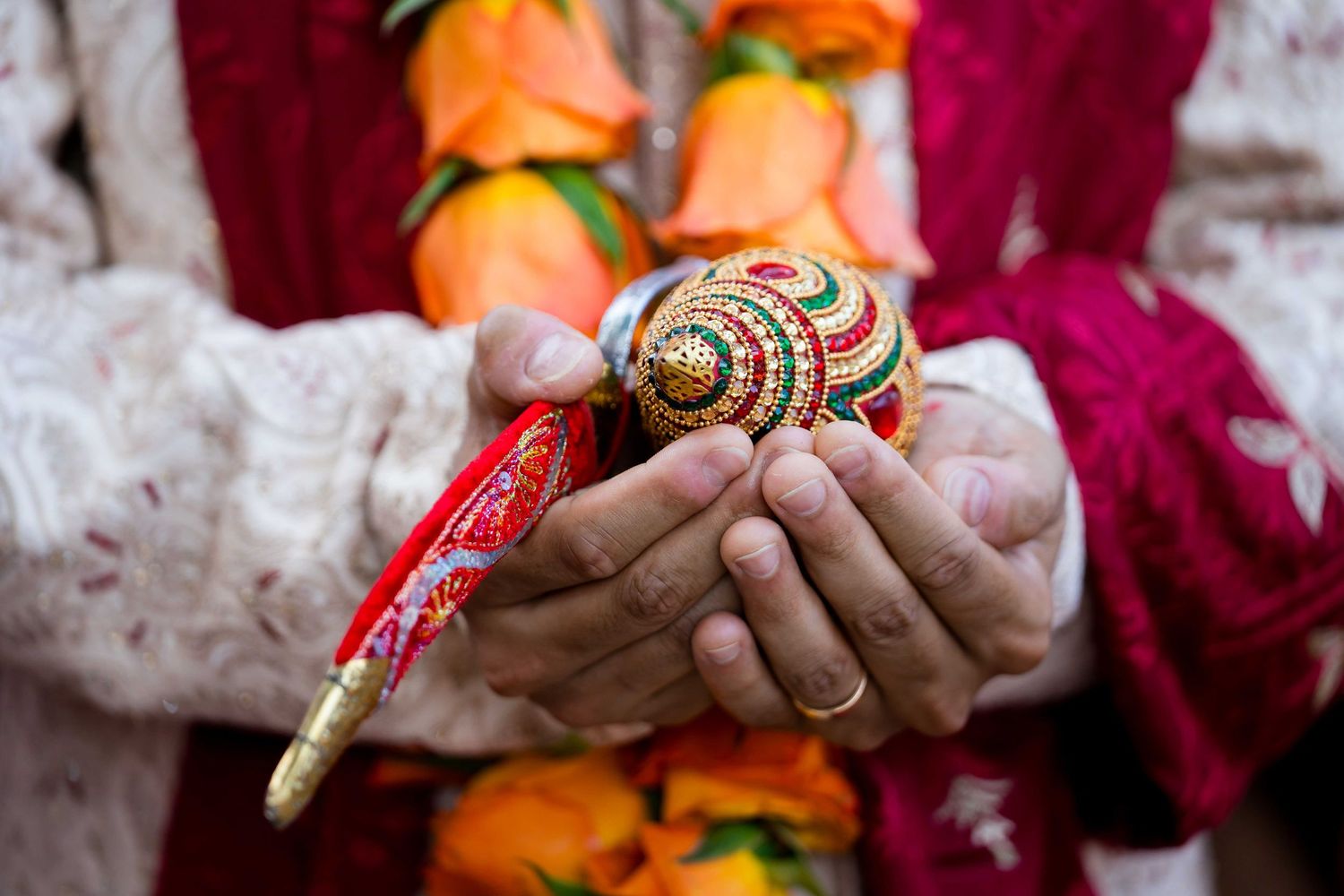 Hands holding traditional Indian wedding ceremony items with orange flowers and red decorative elements in the background.