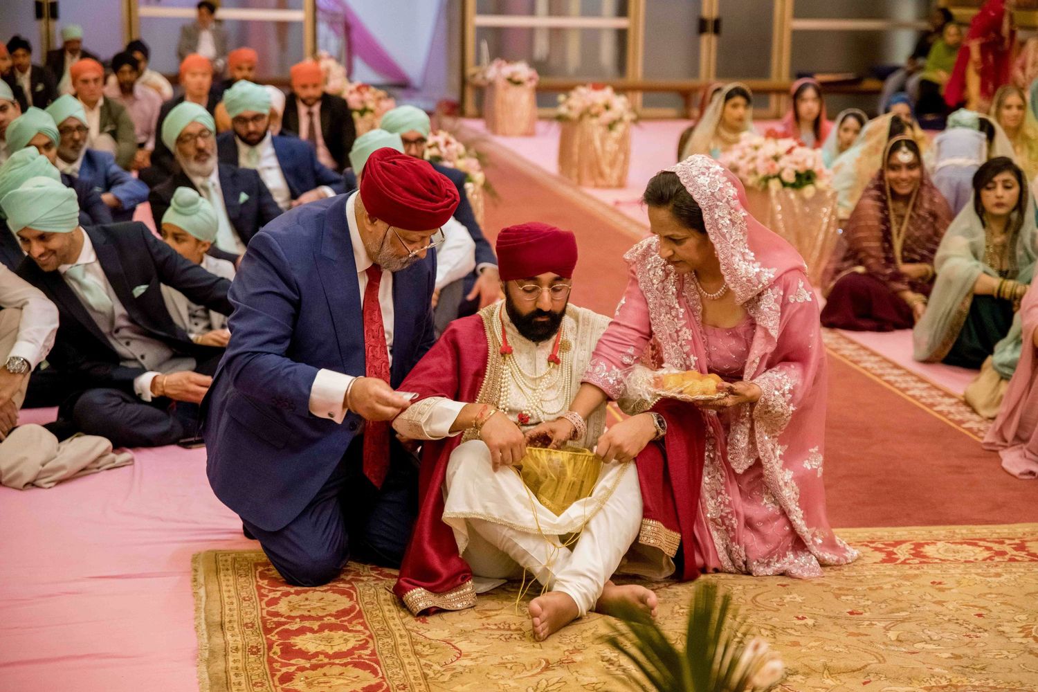 Traditional Sikh wedding ceremony showing ceremonial rituals being performed on red carpeted floor at temple.