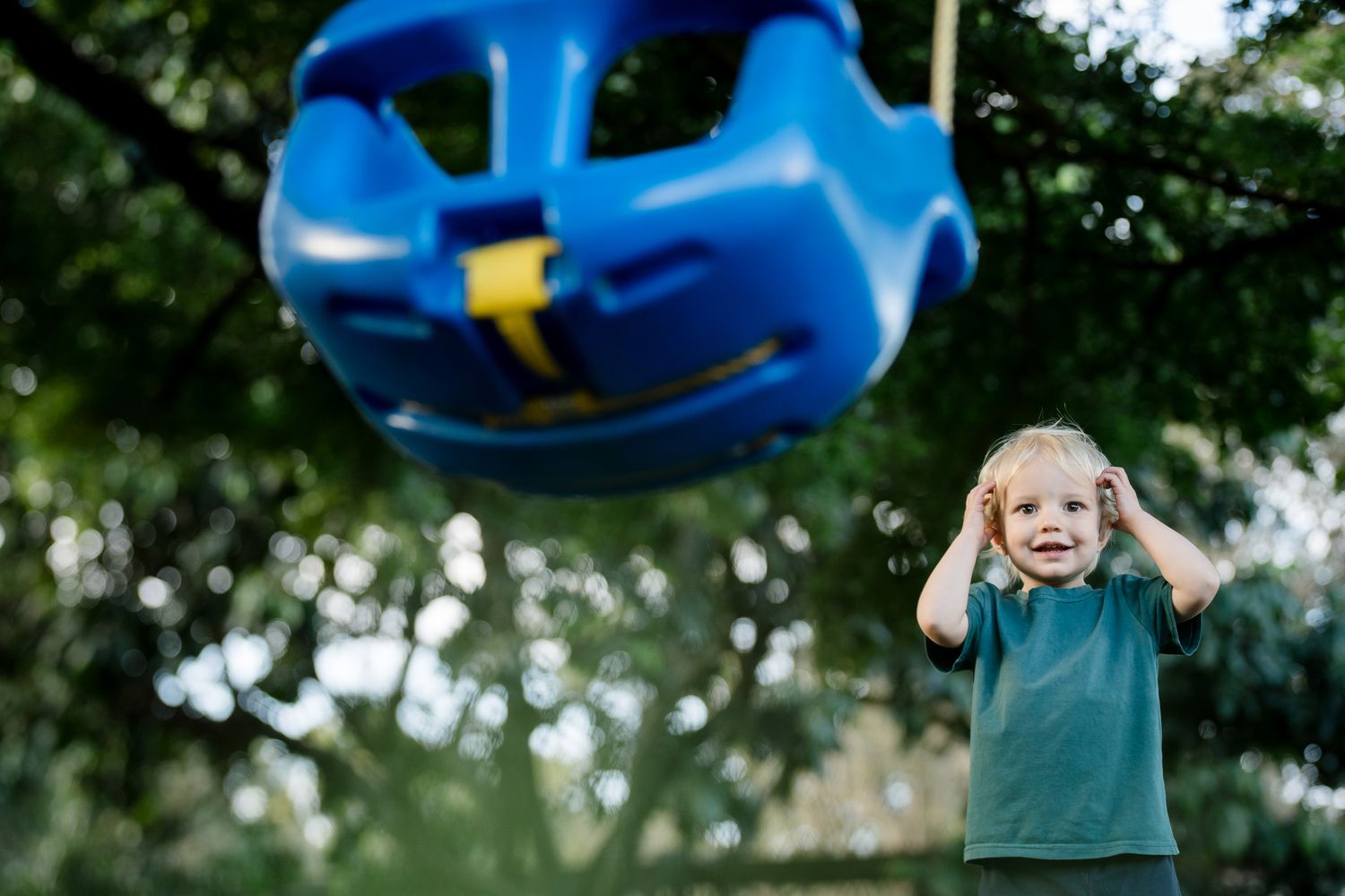 A blue plastic baby swing suspended in mid-air at a playground with a child in a green shirt in the background.