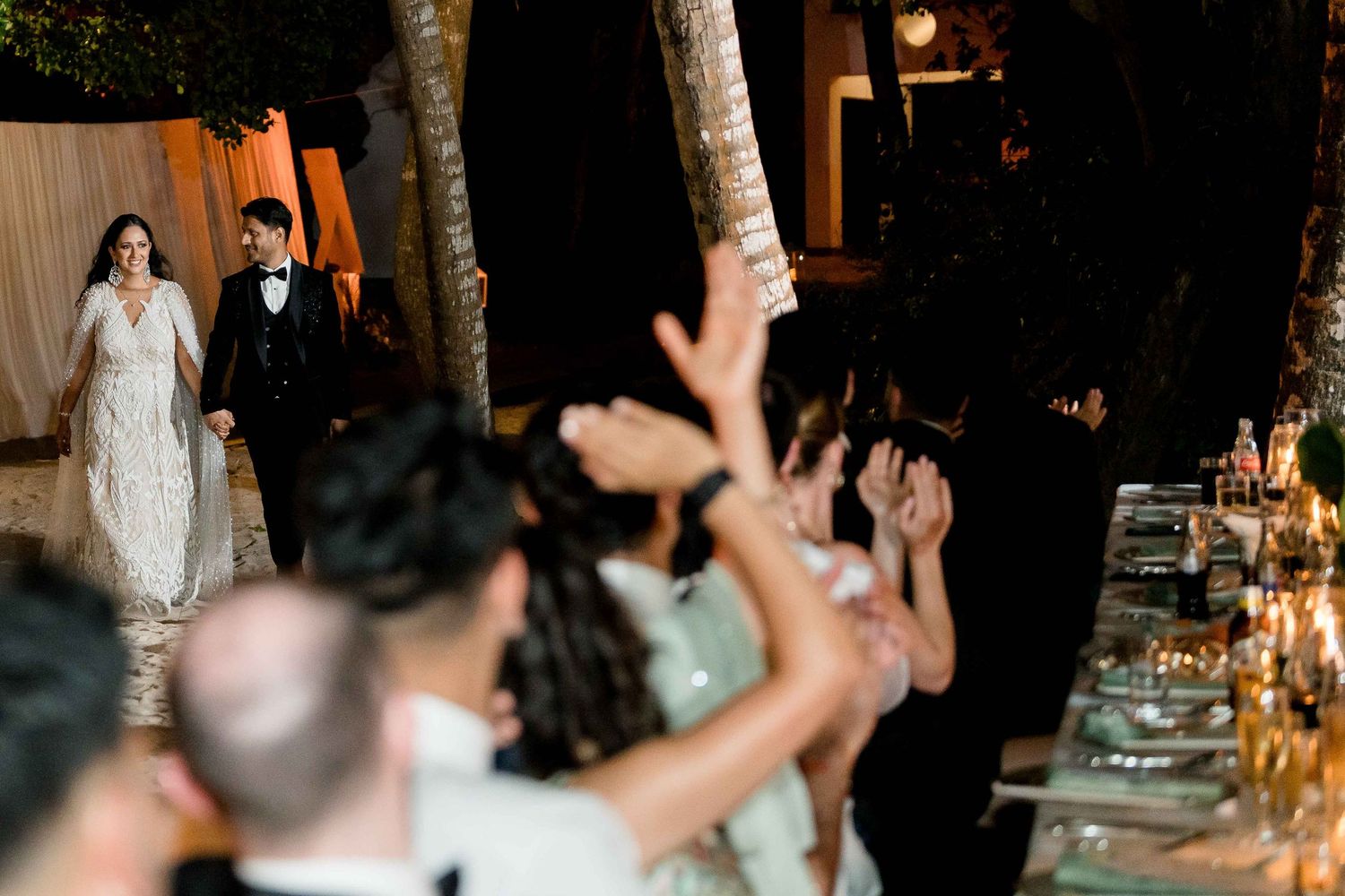 Wedding guests raise their hands in celebration during an evening reception at an elegantly decorated venue.