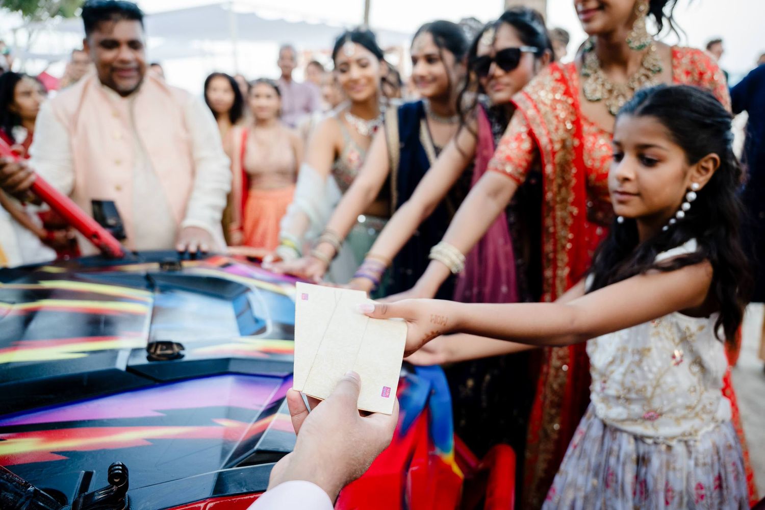 A group of people gather around a car during what appears to be an Indian wedding celebration.