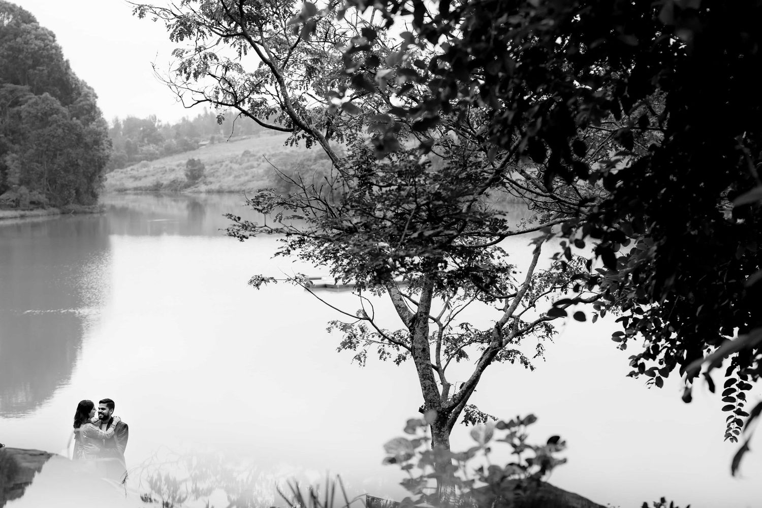 Silhouetted figures sit peacefully by a serene lake surrounded by trees in black and white.