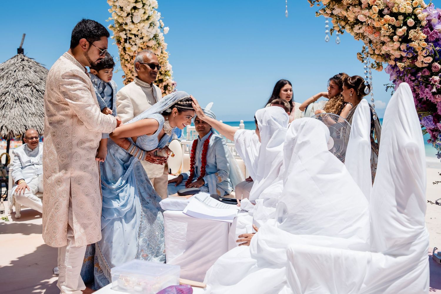 Wedding ceremony guests gathered outdoors under floral decorations on a sunny day with blue skies.