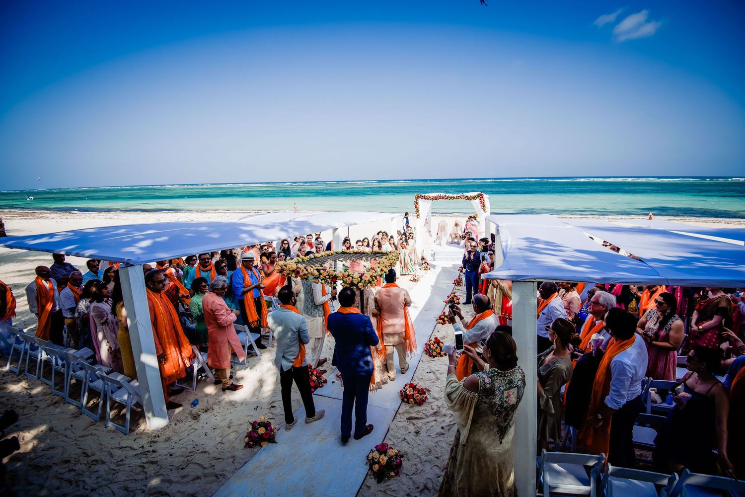 Beach wedding ceremony with guests gathered along the shoreline under bright blue skies.