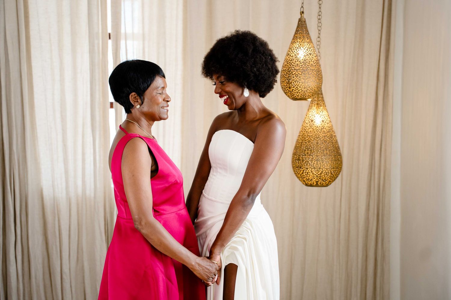 A couple in formal attire embraces romantically in a room with hanging pendant lights and white curtains.