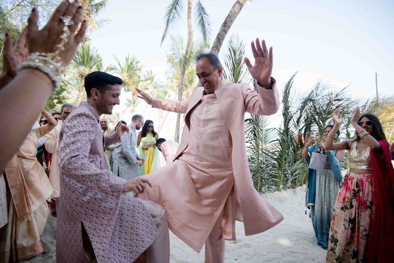 Wedding guests dance and celebrate joyfully on an outdoor patio surrounded by palm trees at a daytime reception.