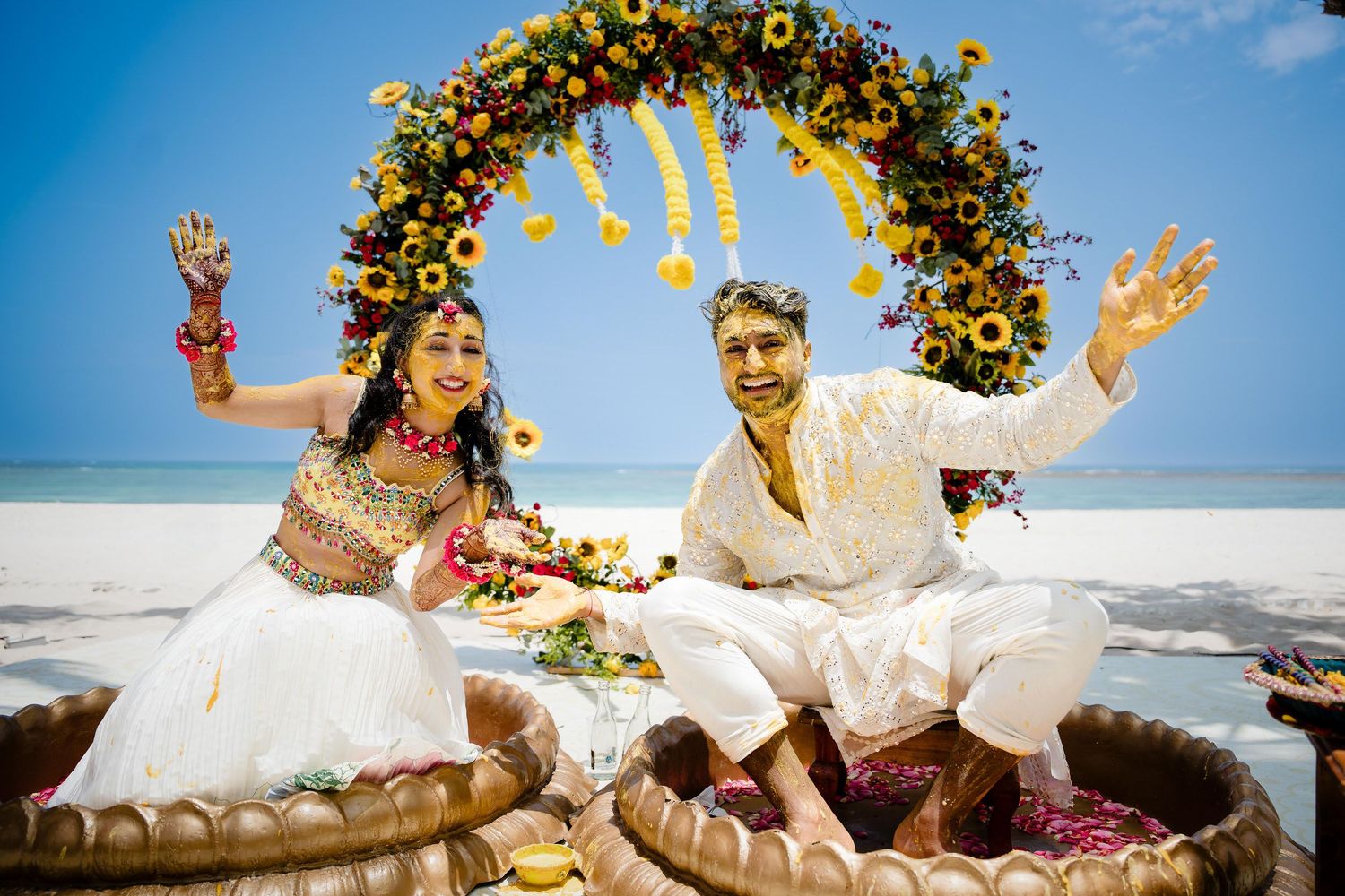 A couple in wedding attire playfully falls back onto a sandy beach with flower petals scattering in the air.