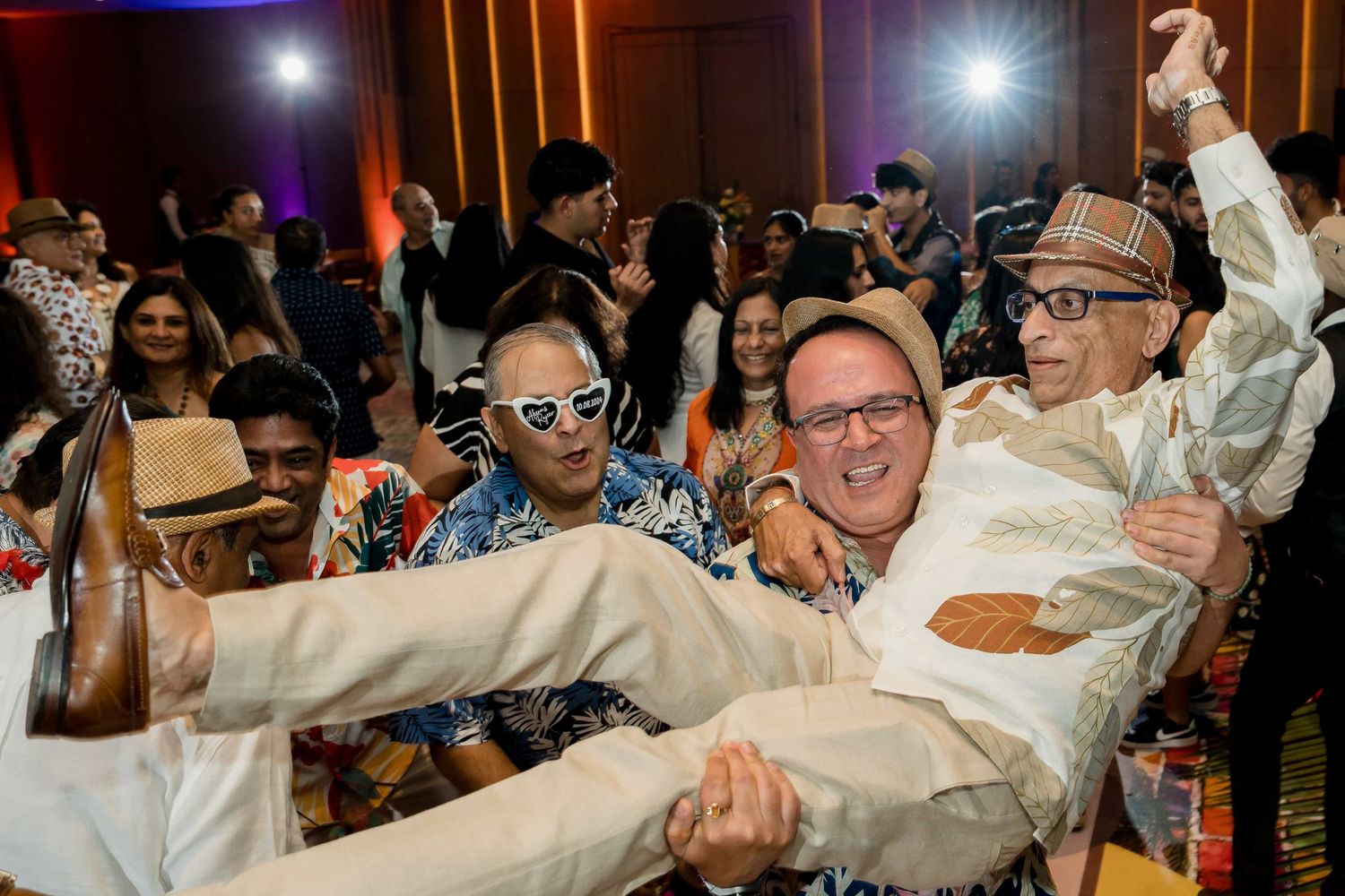 Energetic crowd lifts and carries a person in formal attire during a lively dance party with colorful lighting.