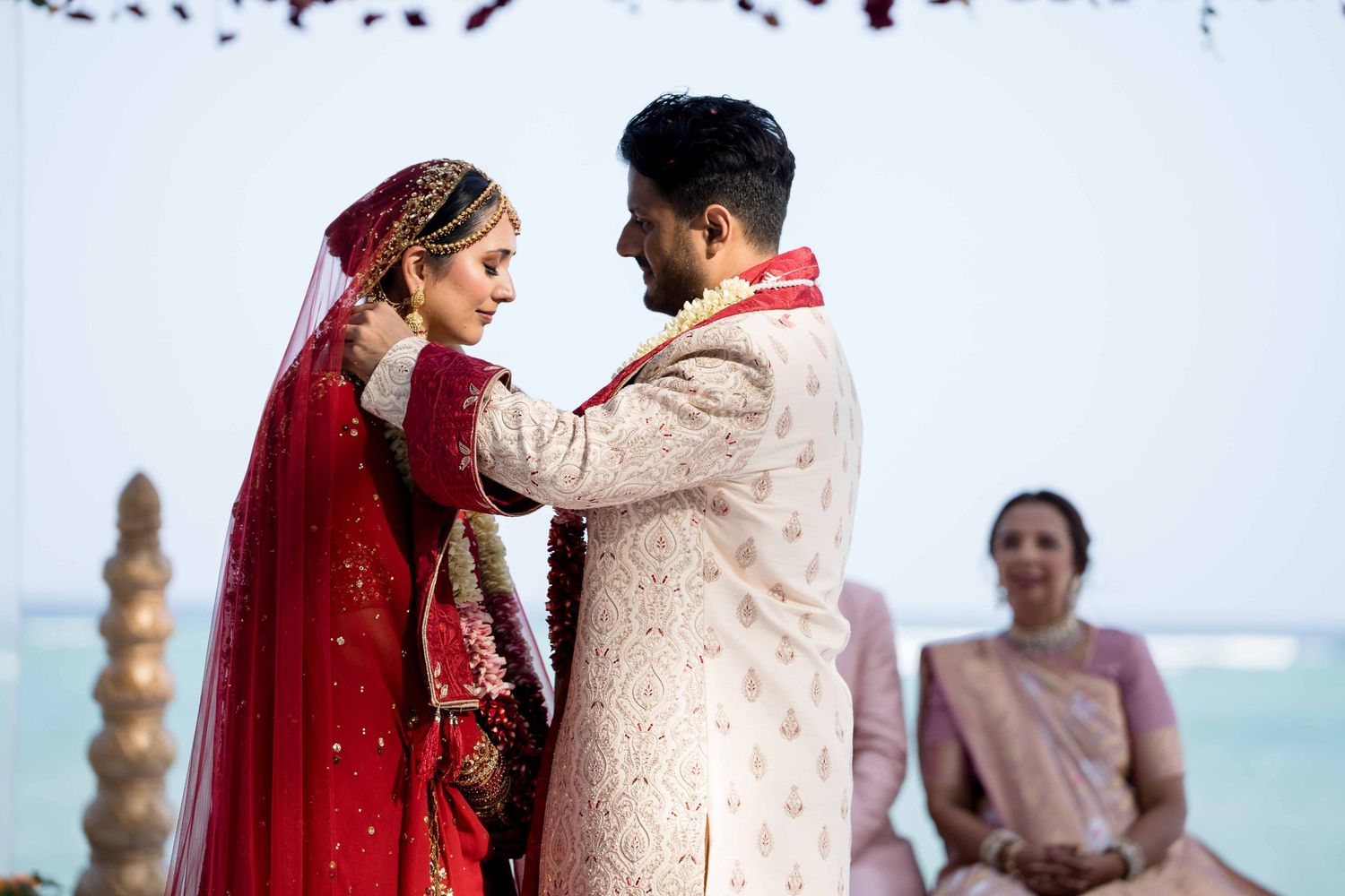Traditional Indian wedding couple in red and cream outfits embrace during their oceanfront ceremony.