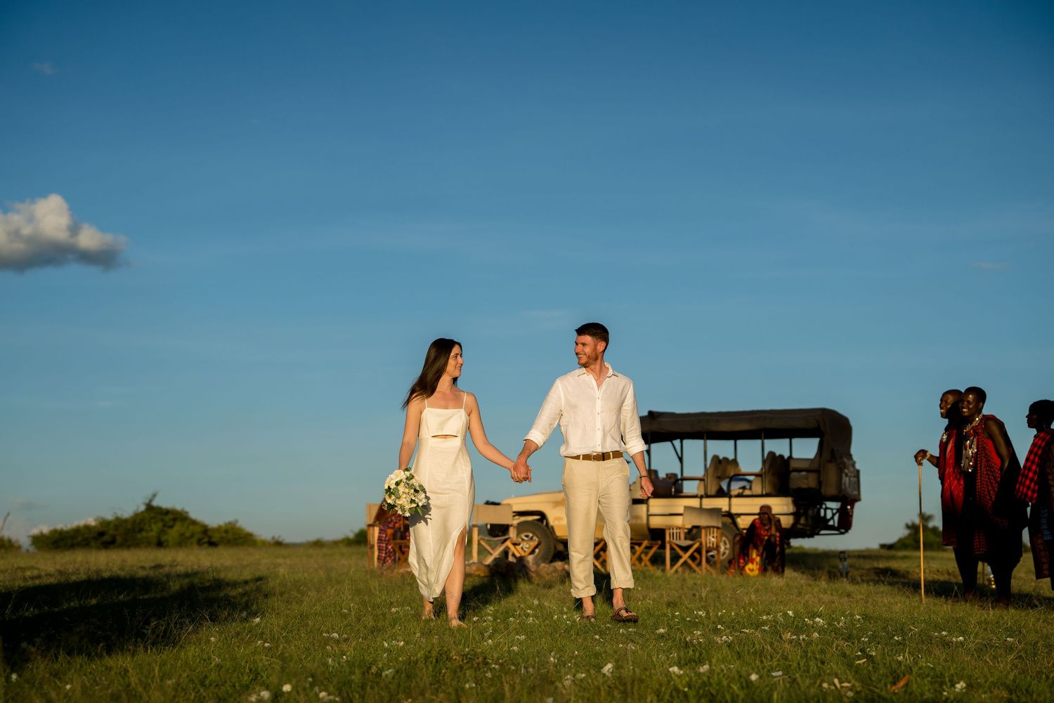 Couple walking hand in hand with safari vehicle in background during golden hour.
