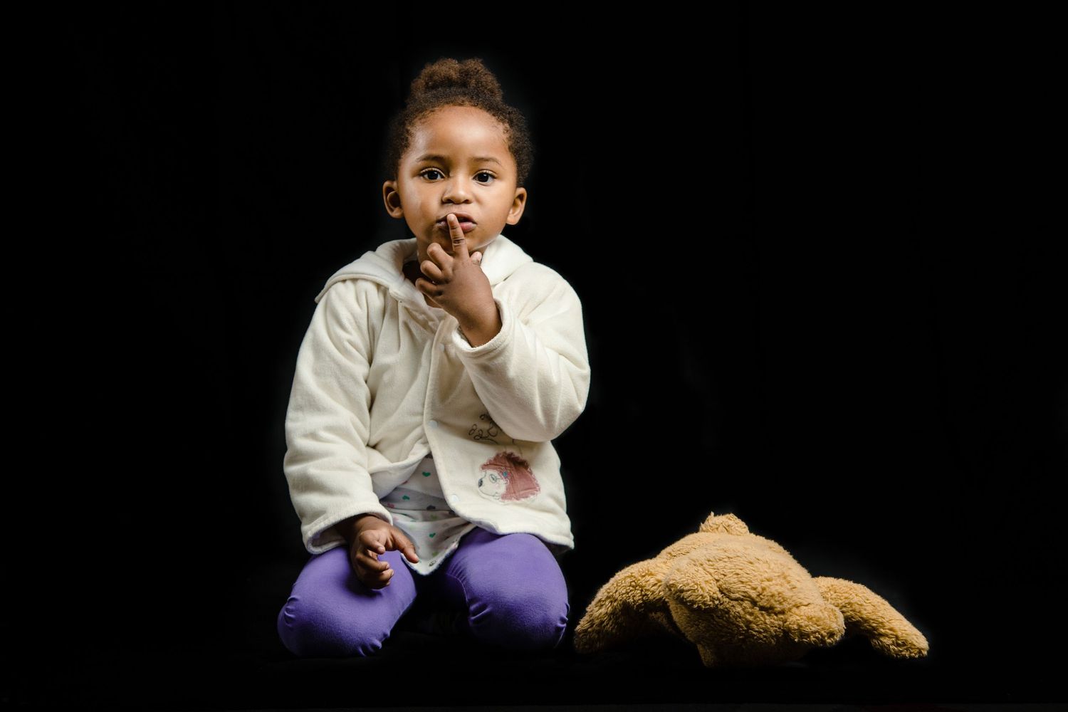 Child sitting with stuffed bear against black background in casual white clothing.