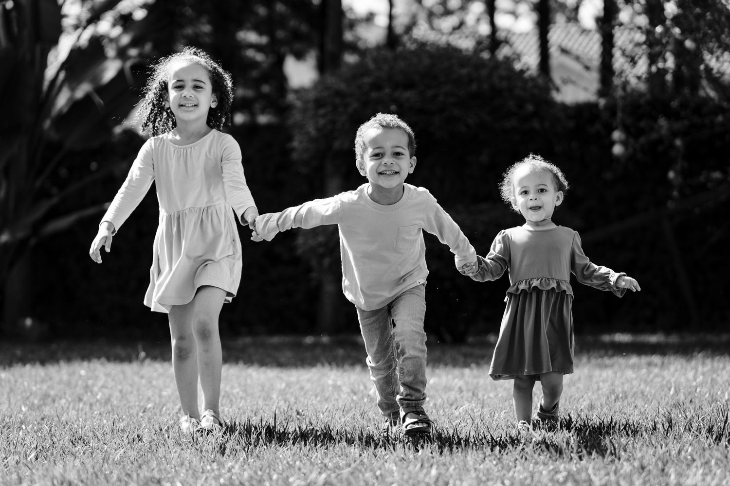 Three children hold hands and run together through a grassy field in a black and white photograph.