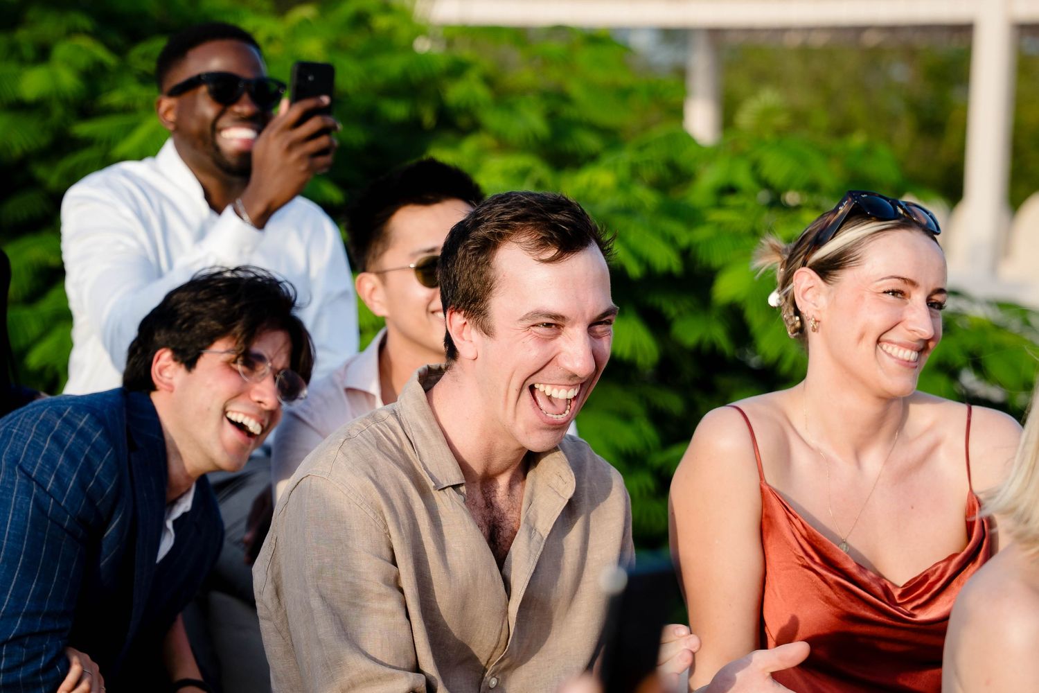 A group of people laugh together while sitting outdoors on a sunny day with green foliage in the background.