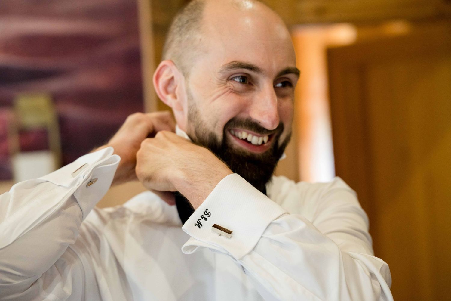 A groom in a black tuxedo laughs joyfully while adjusting his collar during wedding preparations.