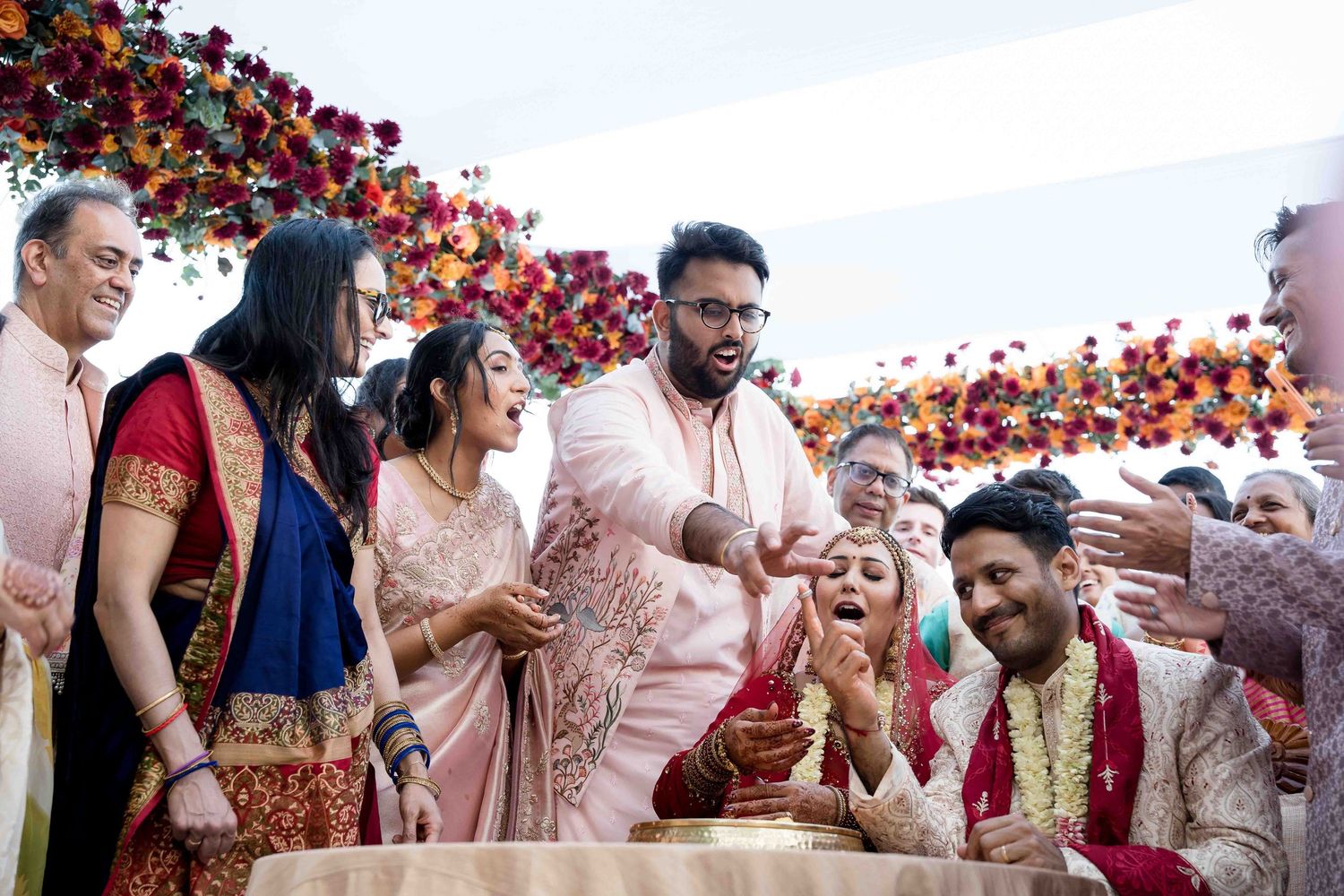 Guests throw red rose petals during an outdoor Indian wedding celebration as the couple walks through the crowd.