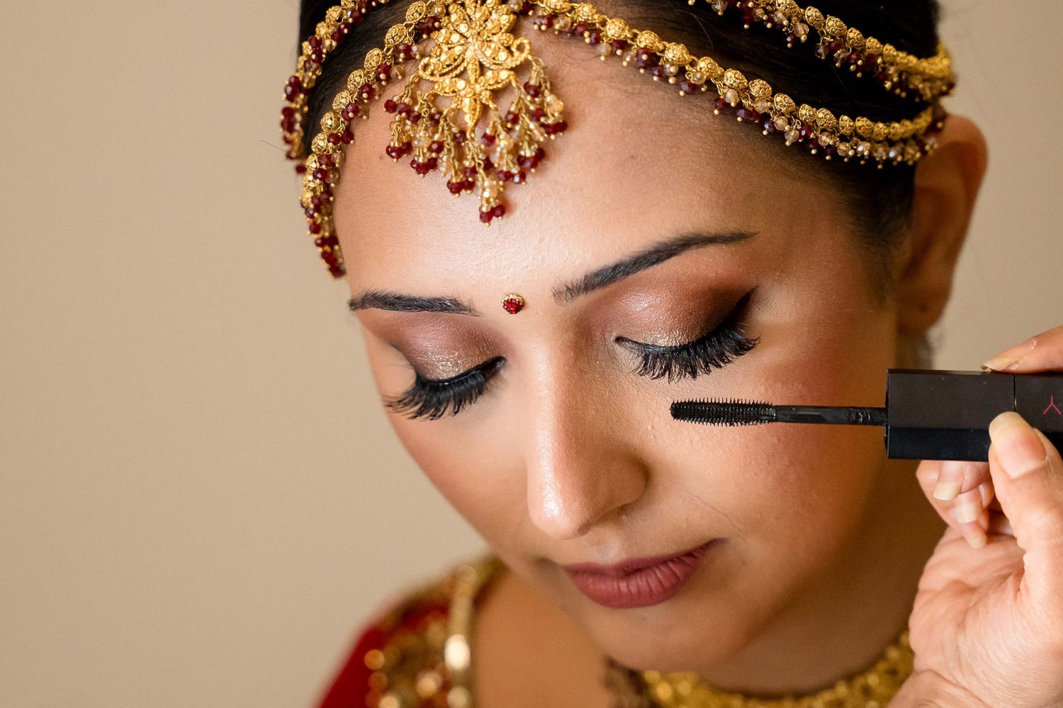 A makeup artist applies mascara to a person wearing traditional Indian bridal jewelry and makeup.