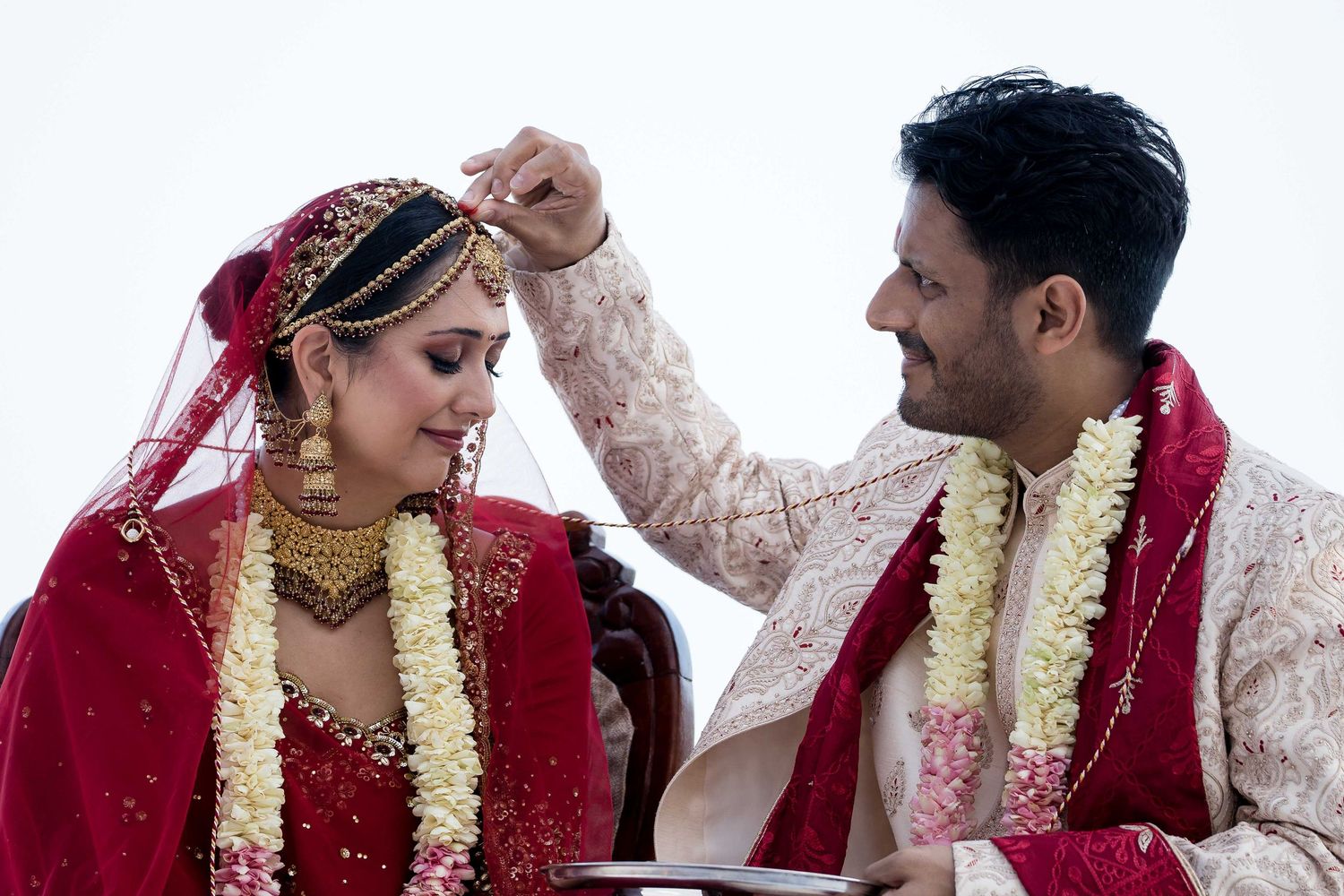 A couple in traditional Indian wedding attire with red and gold garments and floral garlands share a tender moment.