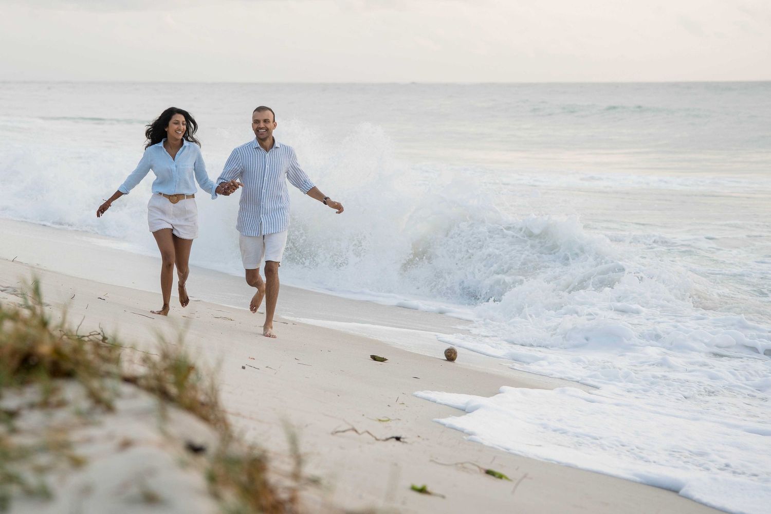 A joyful couple runs together along a sandy beach with crashing waves in the background at sunset.
