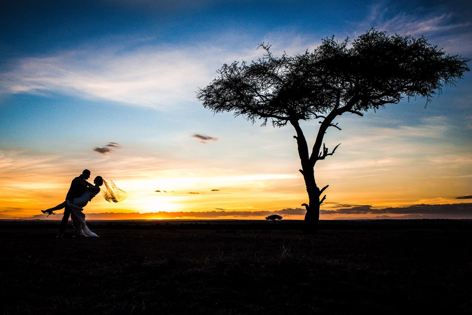 Silhouette of photographer against dramatic sunset sky with lone acacia tree.