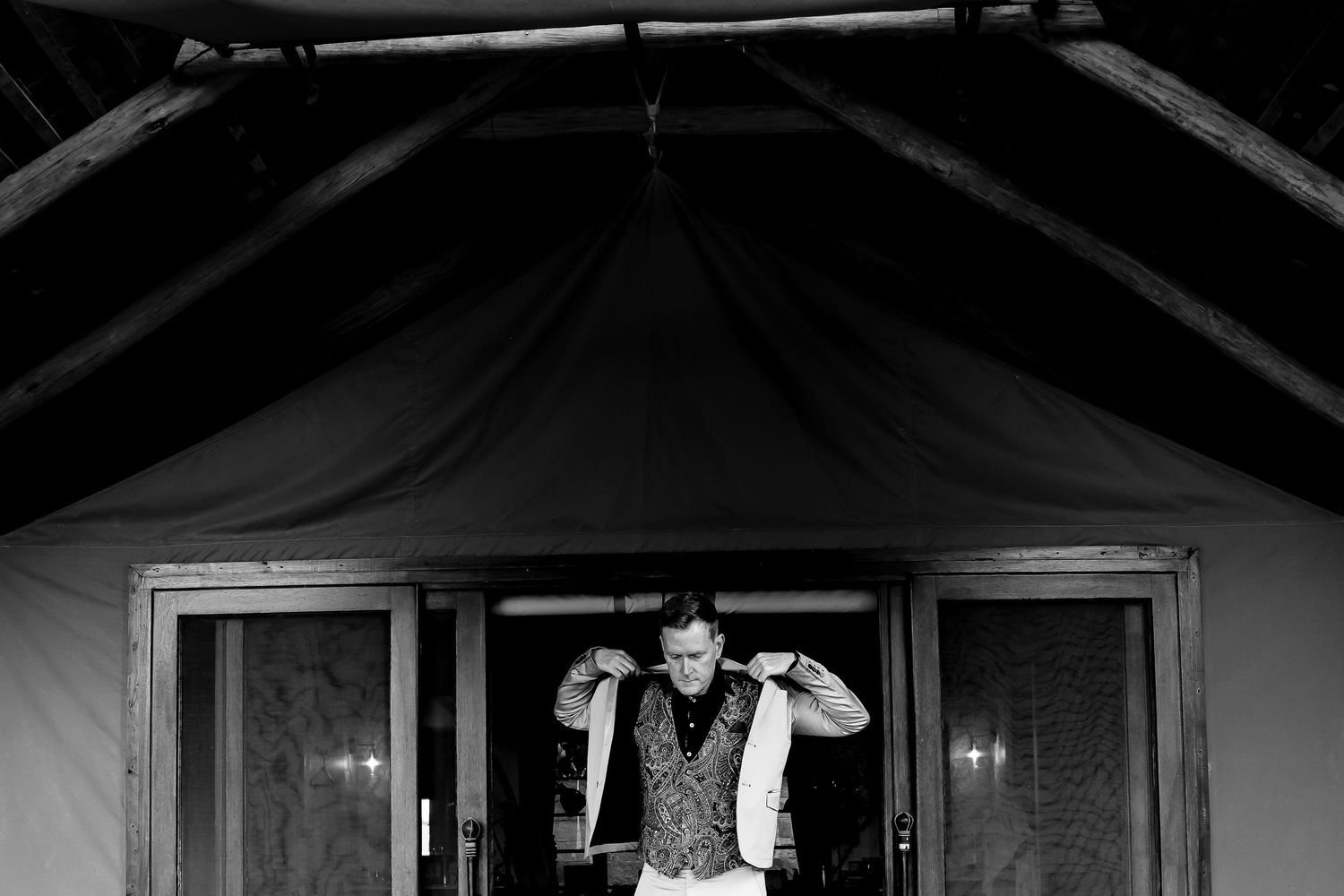 Black and white photo of a tattooed groom adjusting his vest inside a rustic tent venue.