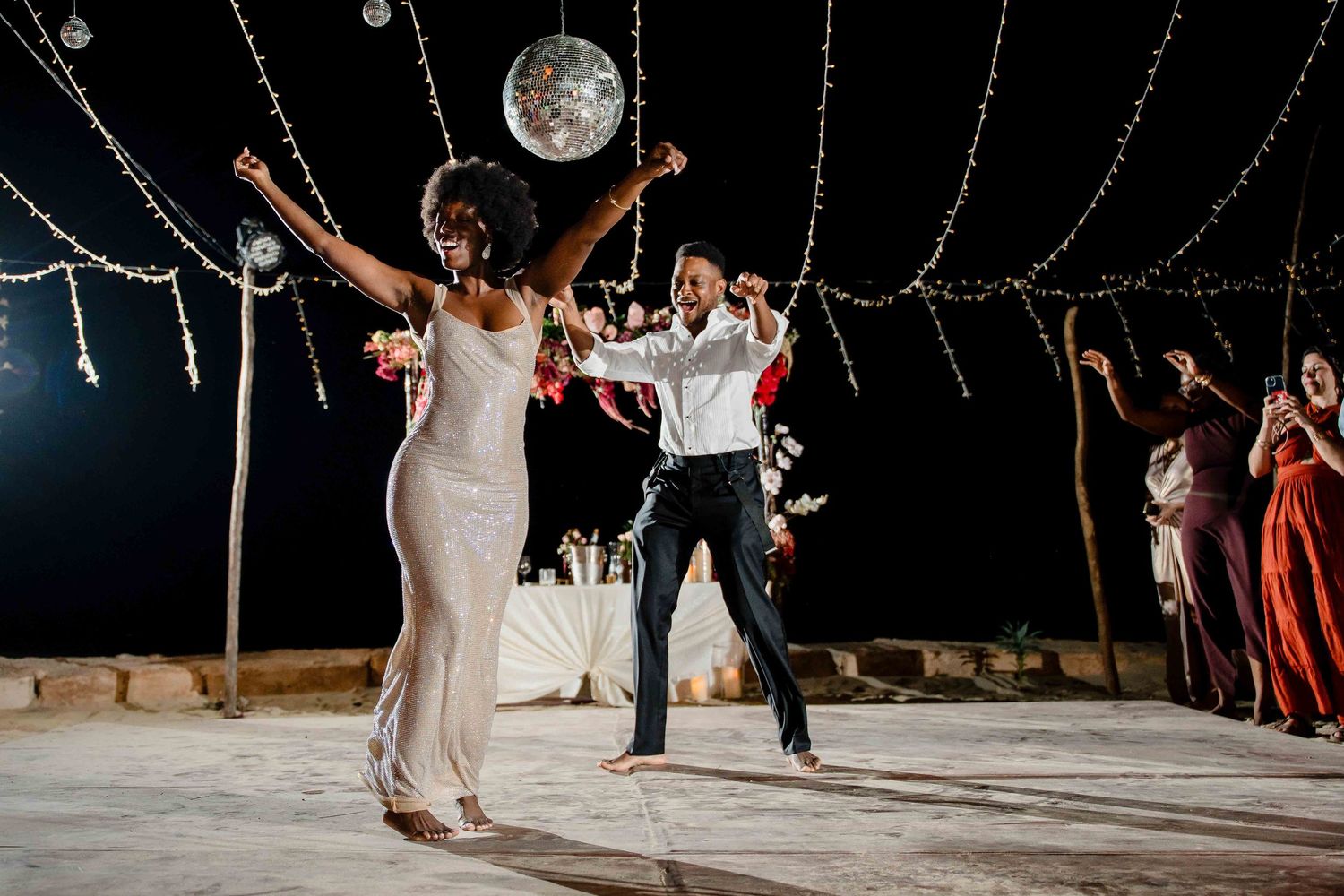 Wedding guests dance and celebrate on an outdoor dance floor beneath twinkling string lights at night.