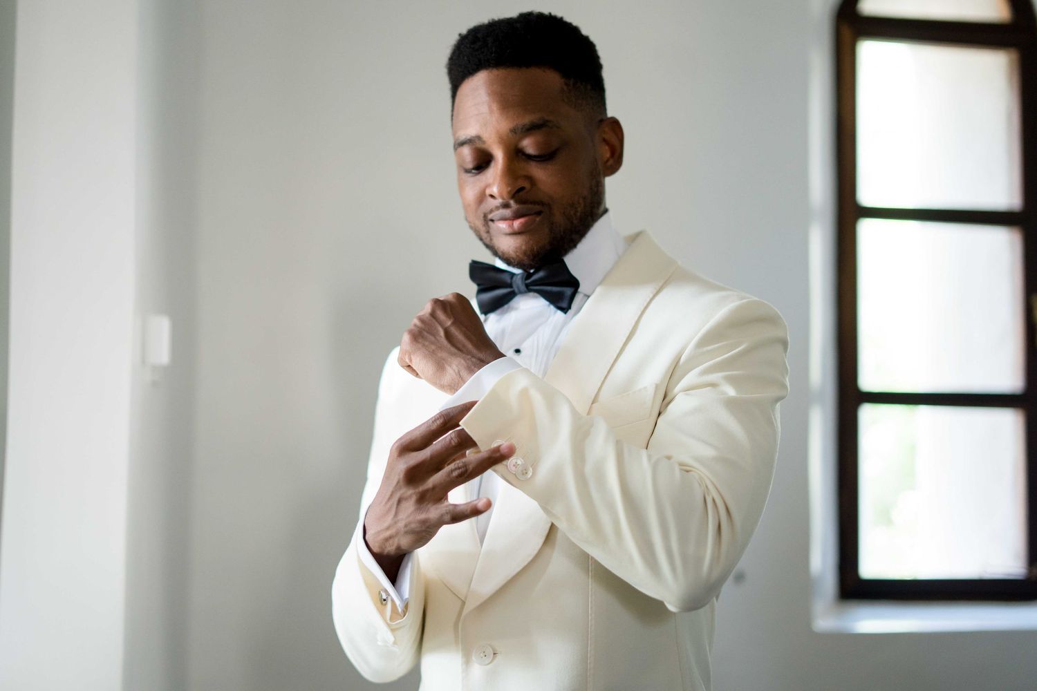 A groom in a cream white tuxedo and bow tie adjusts his cuff while getting ready for his wedding ceremony.