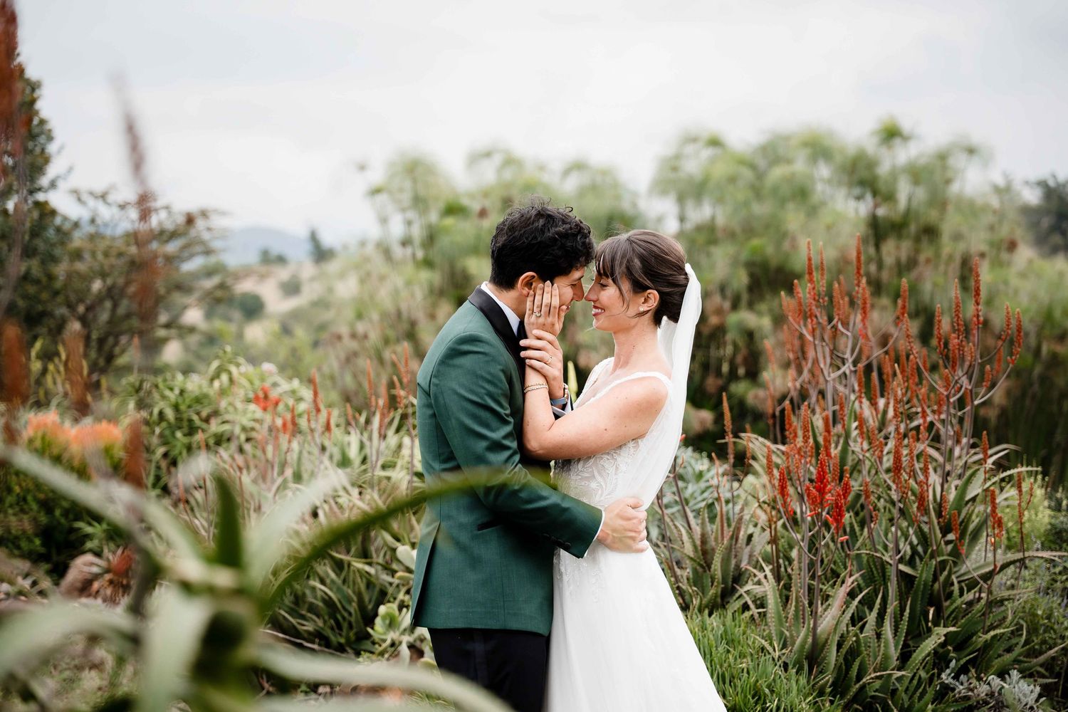 A romantic couple embraces in a garden filled with red flowers and lush greenery during a wedding photoshoot.