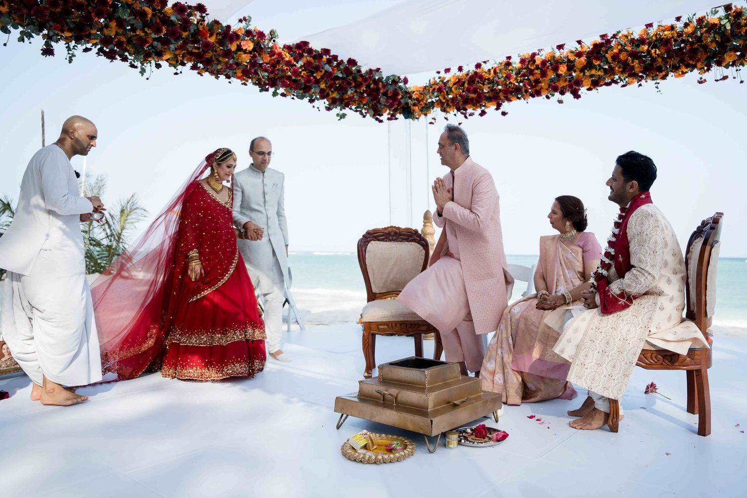 Traditional Indian wedding ceremony with participants seated on chairs under a floral canopy during a beachside ritual.