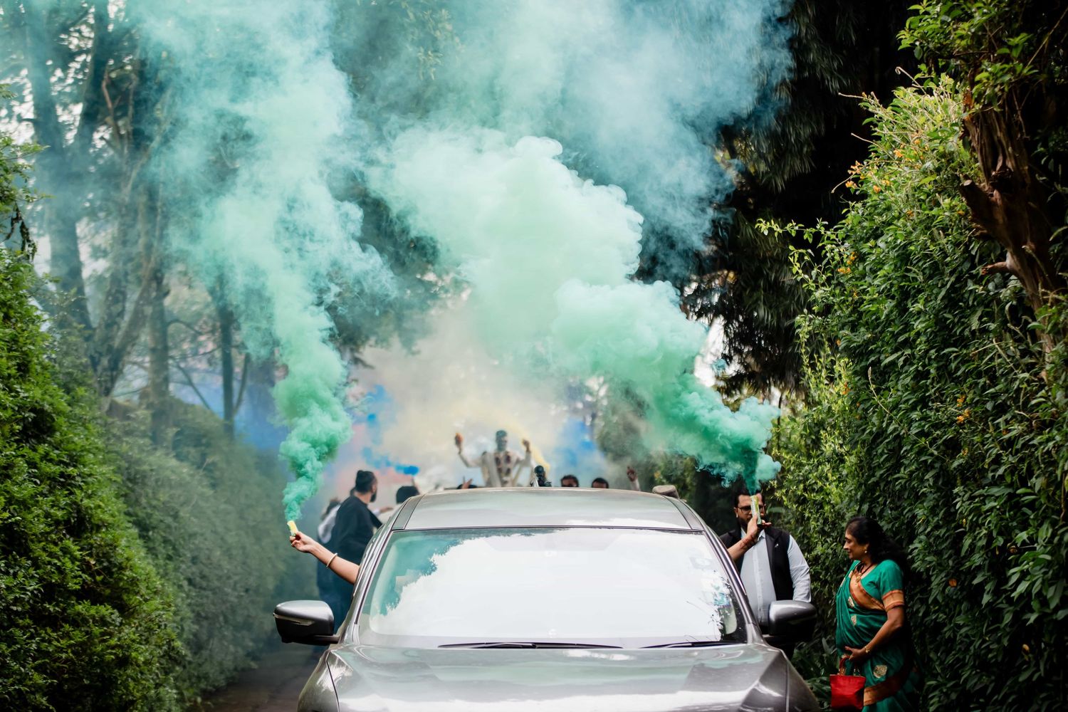 Celebratory scene with turquoise smoke bombs around a car surrounded by greenery.