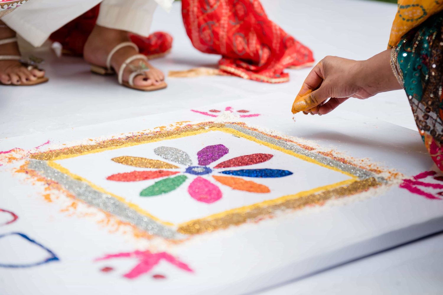 Colorful rangoli art design being created with flower petals and glitter on white surface.