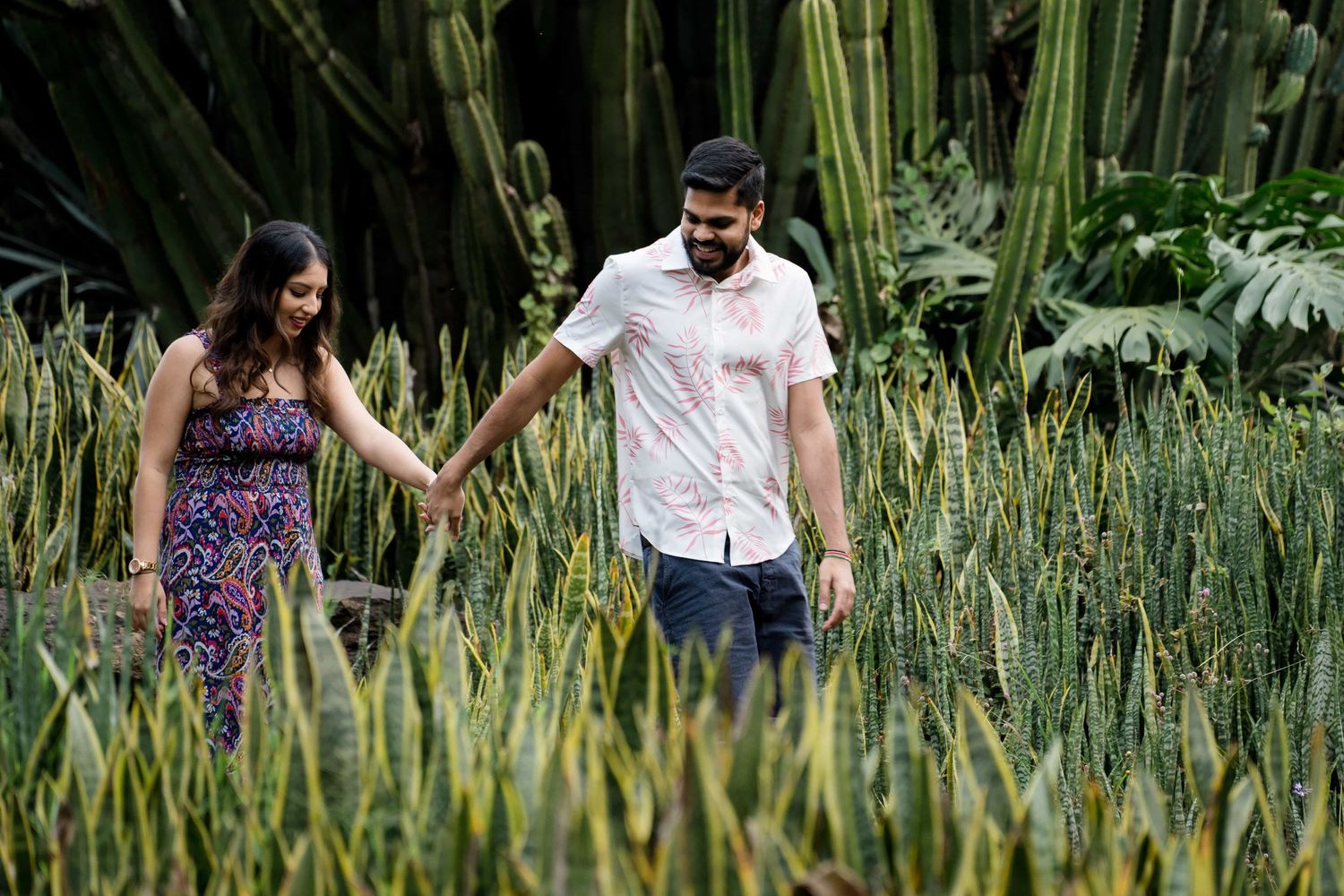 A couple walks hand in hand through tall green grasses in a lush garden setting.