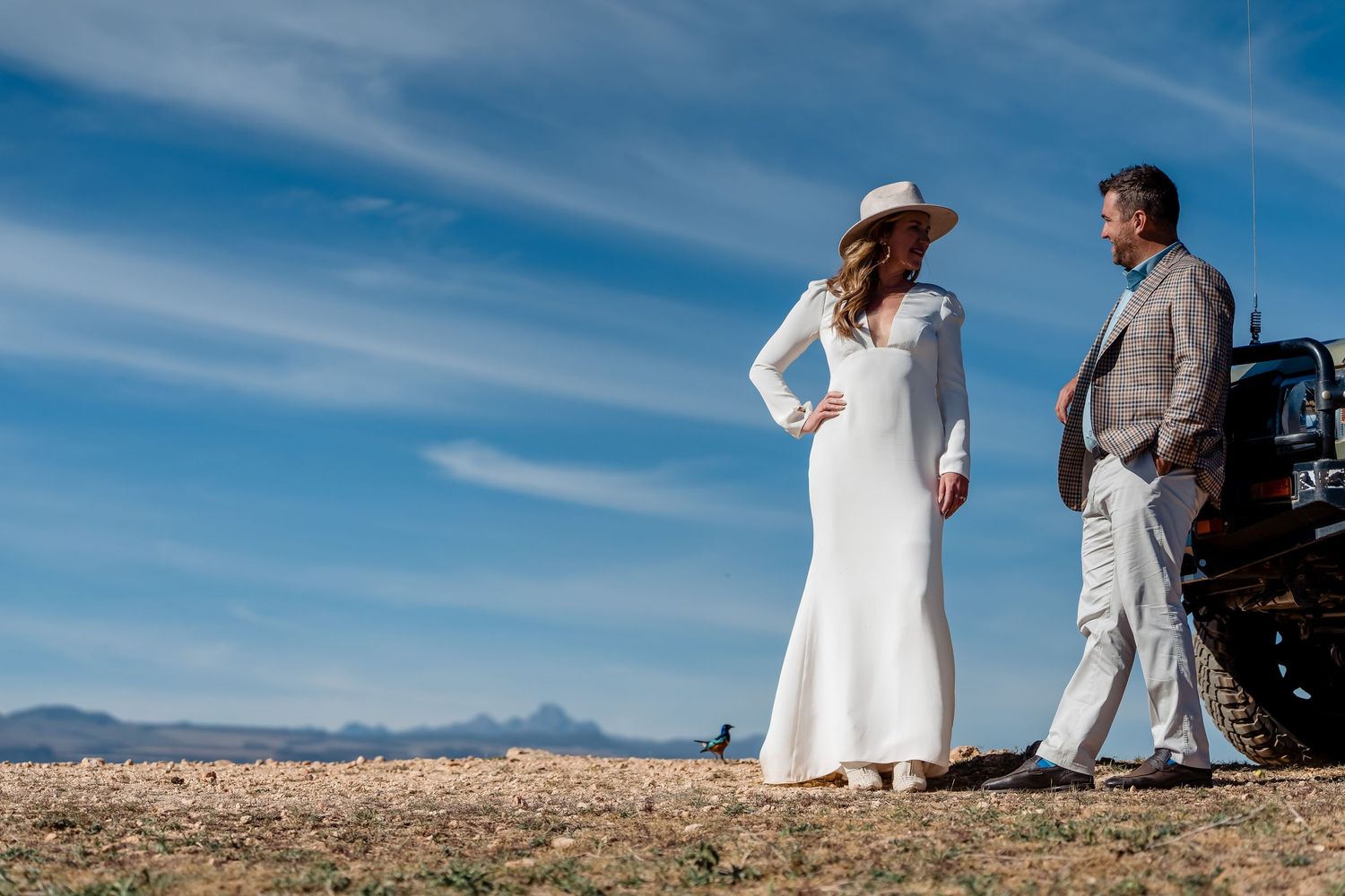 A couple in white and grey formal wear stands on a hilltop against mountains and blue sky.