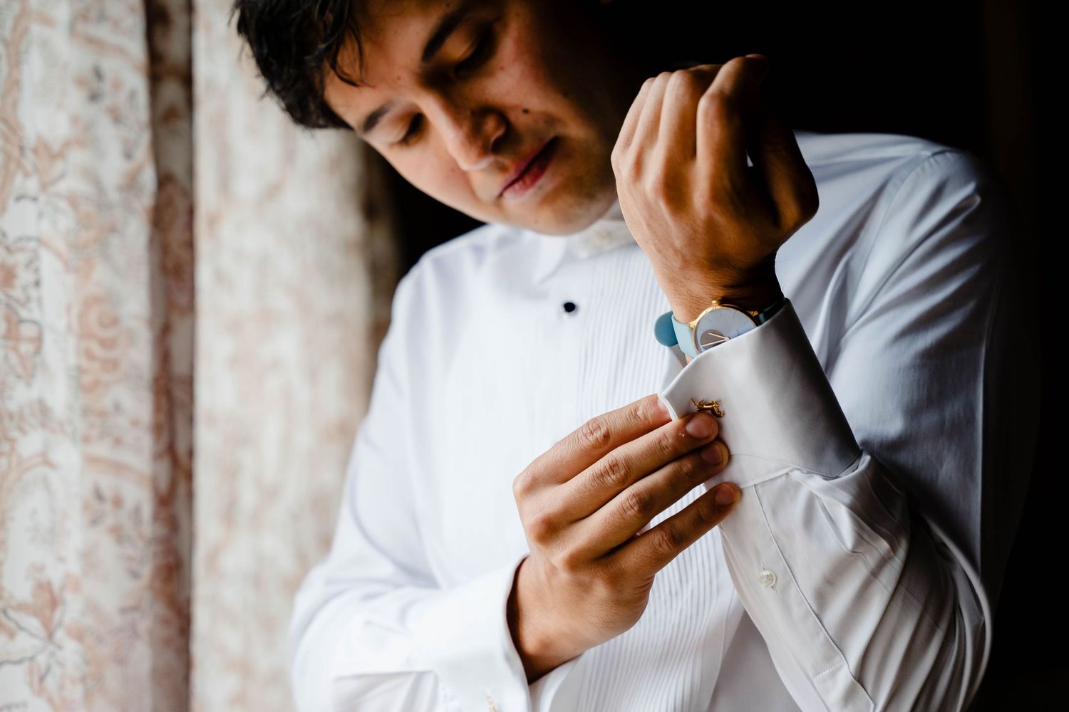 Professional in white shirt and dark blazer leans against wall while looking down thoughtfully in studio setting.