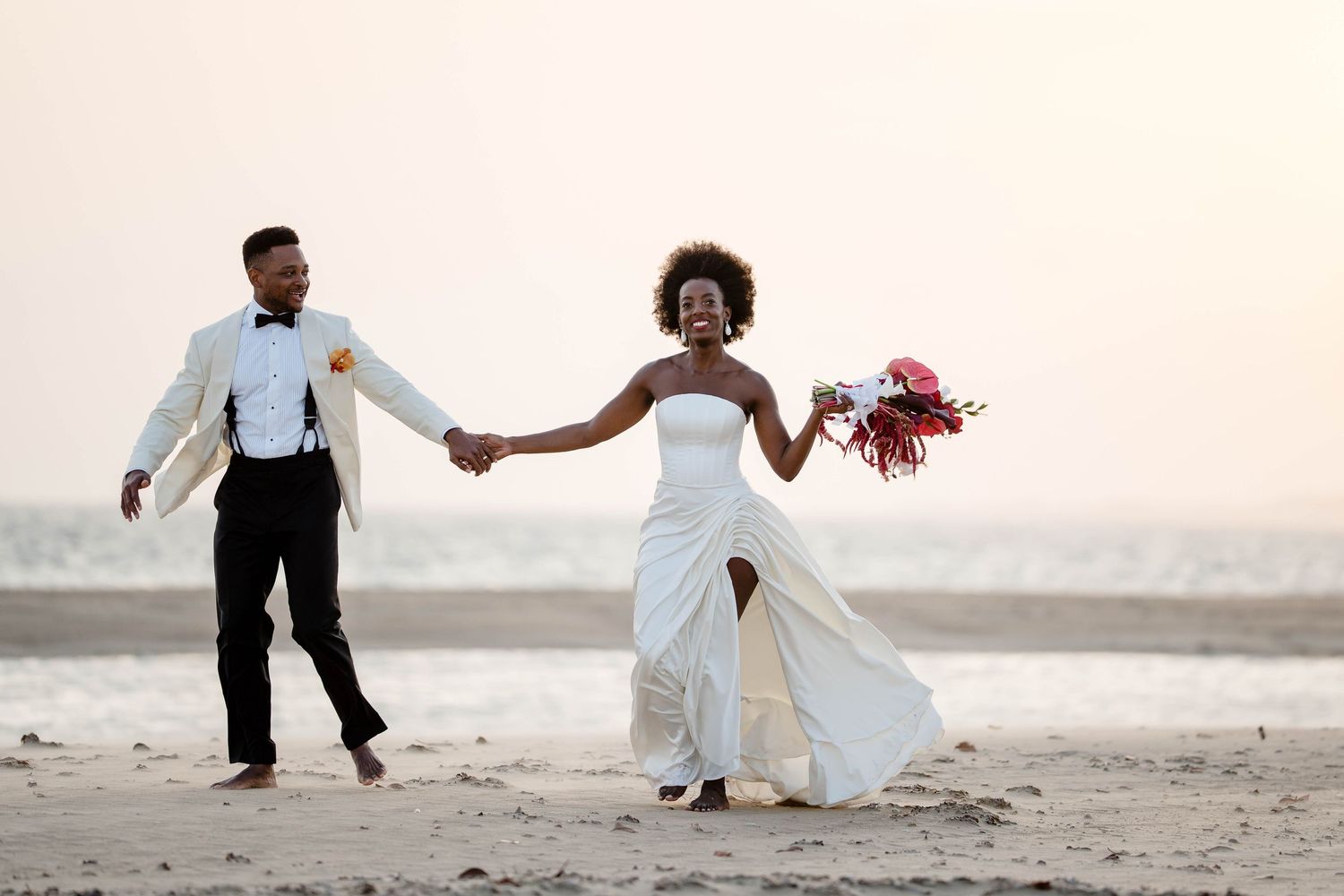 A joyful couple in wedding attire runs along a sandy beach at sunset holding hands as the bride carries a red bouquet.