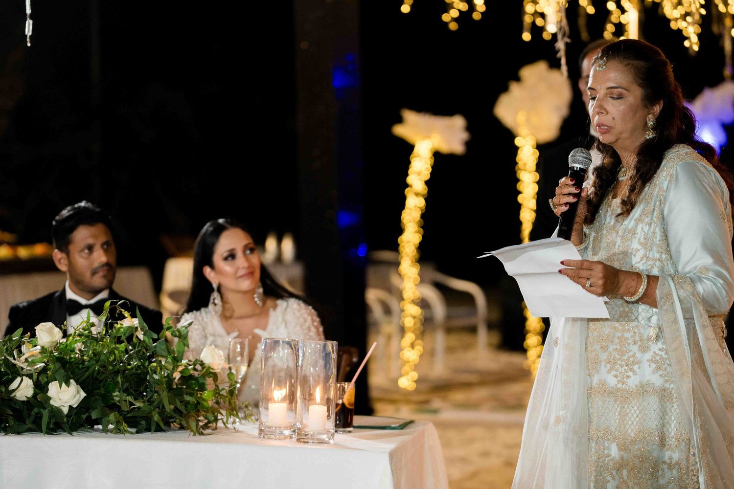 Wedding reception guests seated at tables with elegant decor and warm lighting in an evening celebration setting.