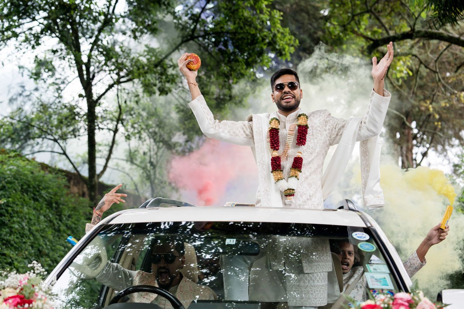 A traditional Indian wedding celebration with smoke bombs creating vibrant colors around a decorated car.