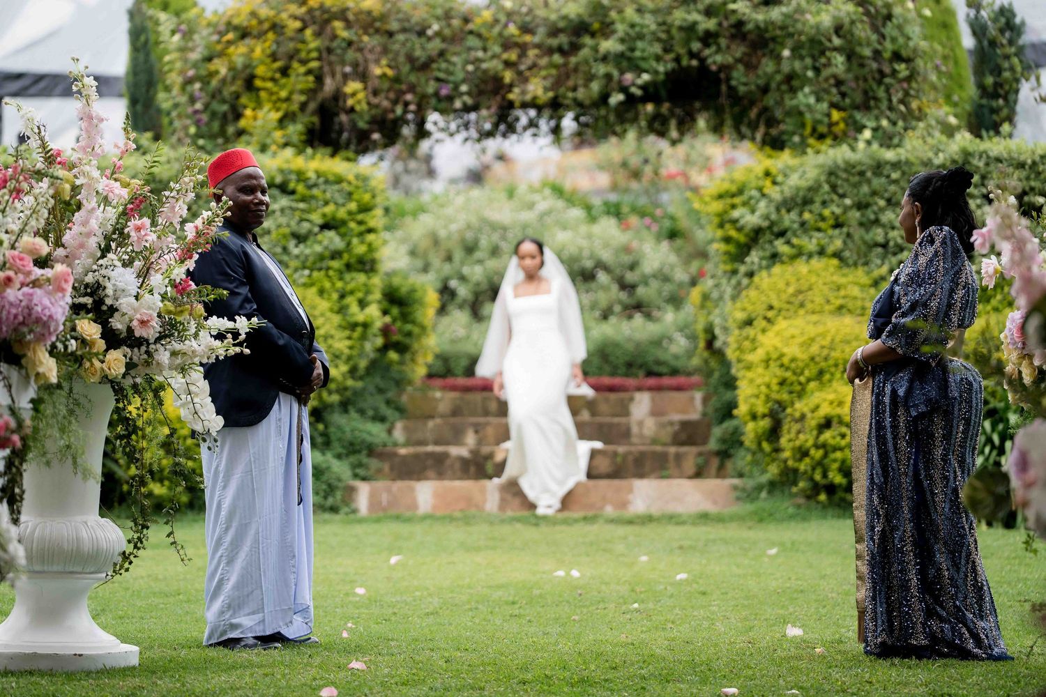 Wedding party members in formal attire walk across a lush green lawn during an outdoor garden ceremony.
