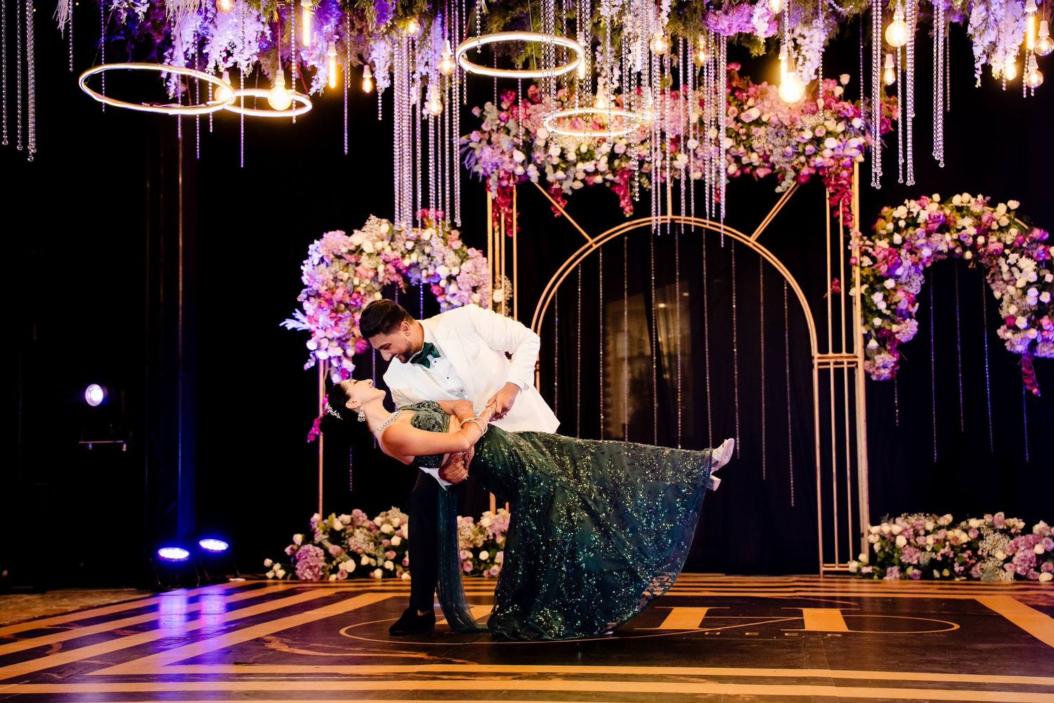 A couple performs an elegant dance dip on a dark dance floor beneath hanging lights and floral arches.