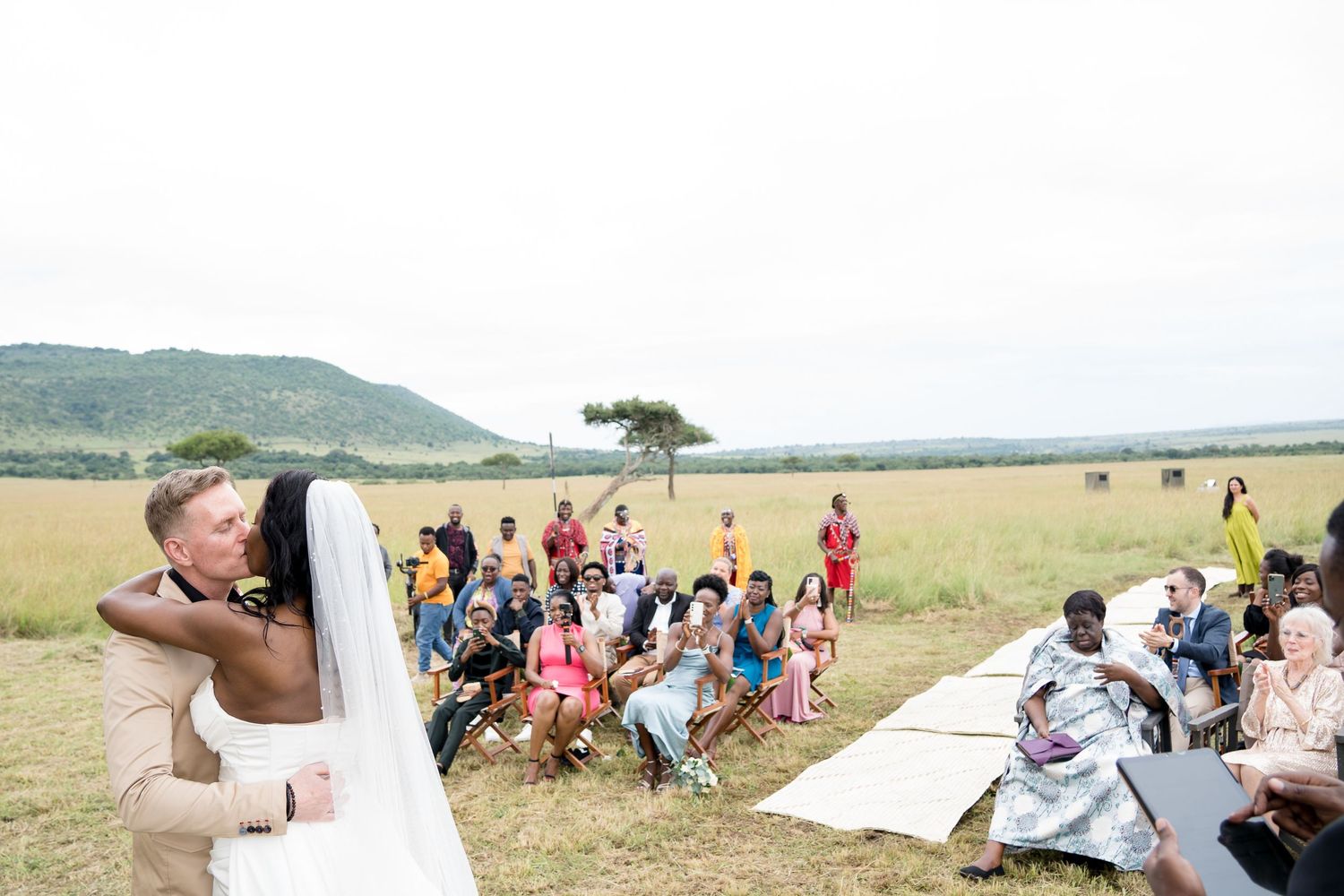 Wedding celebration in an outdoor African savanna setting with guests gathered under an acacia tree.