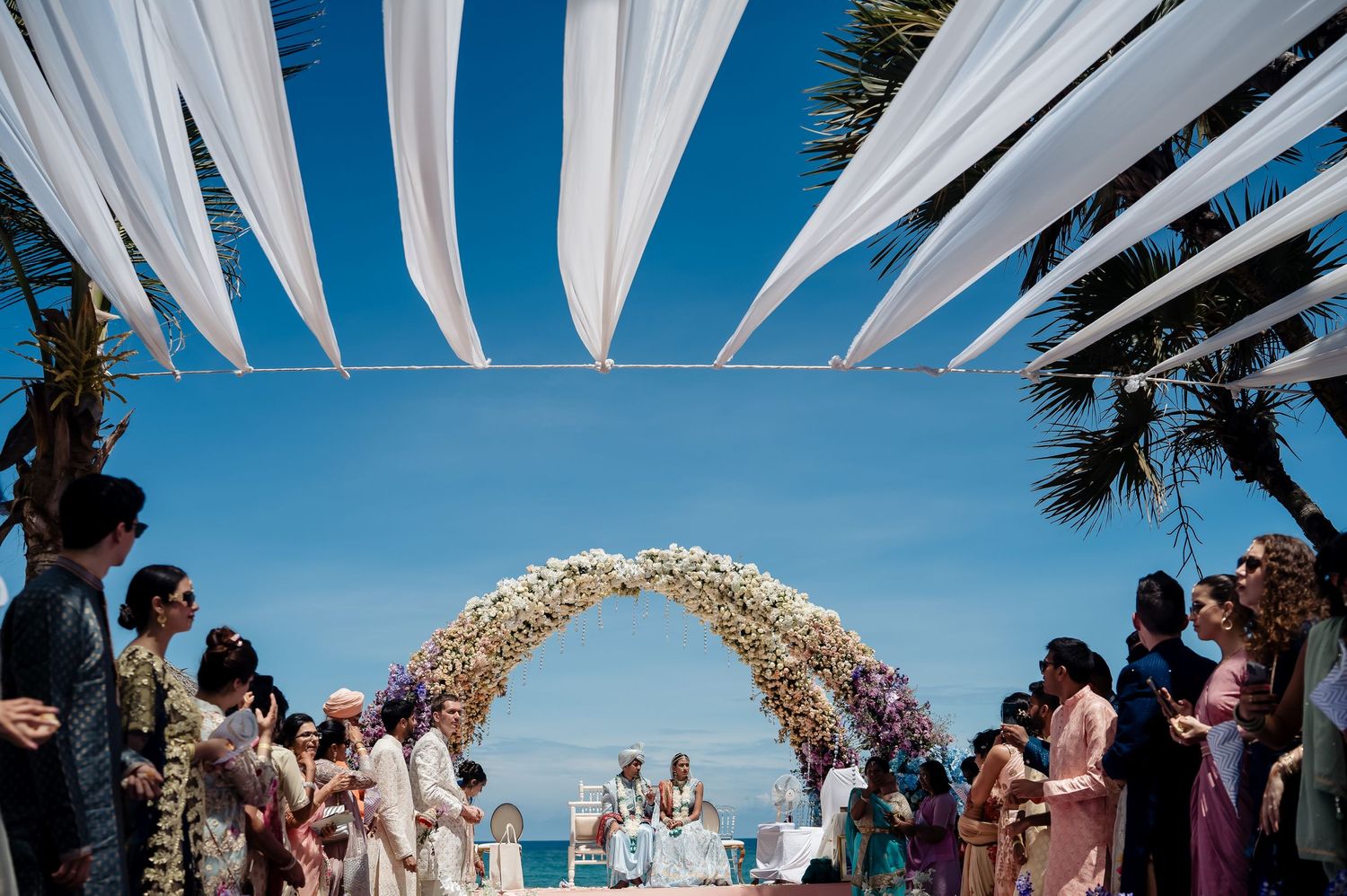 Beach wedding ceremony setup with white fabric canopy and floral arch overlooking the ocean.