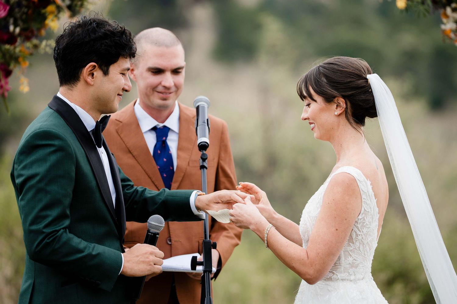 A couple exchanges wedding vows and rings during an outdoor ceremony with a rustic wooden backdrop.