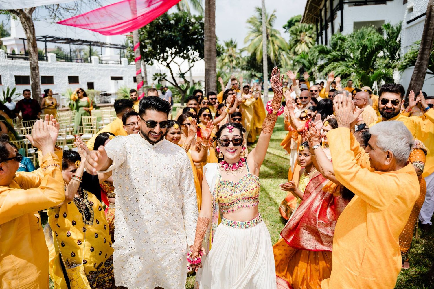 Joyful wedding guests celebrate during an outdoor Indian ceremony with yellow and pink decorations.