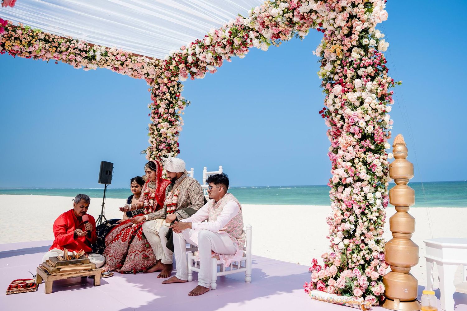 A beachfront wedding ceremony setup with pink floral arches and wooden benches against the turquoise ocean backdrop.