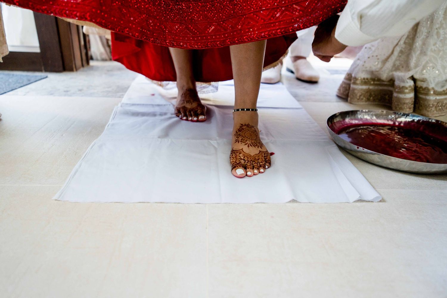 Decorated feet with henna stand on white cloth near a red dress hem.